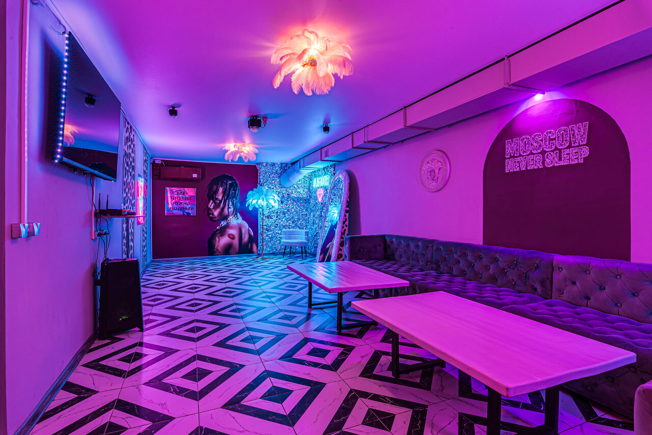 Арендовать Неоновый лофт Гламур. Лофт-пространство с узорчатым полом, фиолетовым и синим неоновым освещением. На стенах постеры, неоновые надписи, флористический декор. Длинный темный диван вдоль стены, светлые прямоугольные столы. Надпись "Moscow Never Sleep" на стене.