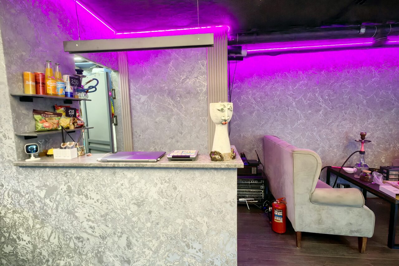Арендовать Зал с кухней площадью 95 м². Помещение с барной стойкой и зоной отдыха. Стойка выполнена из серого камня, за ней на полках напитки и закуски. Мягкий диван и столик справа. Фиолетовая подсветка на потолке. Серые стены с текстурой. Пожарный кран и кальян на столике.