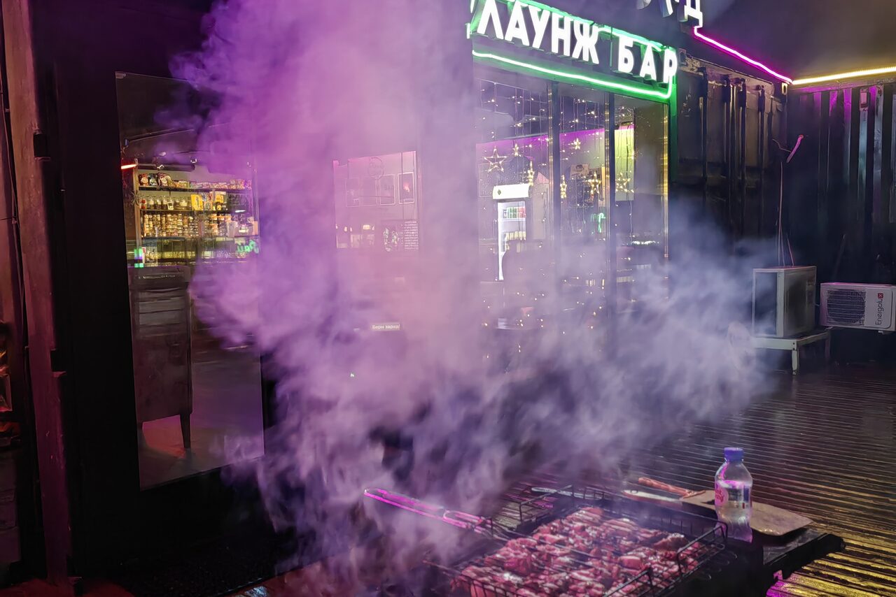 Арендовать Зал с кухней площадью 95 м². Лаунж-бар с неоновыми вывесками, столиками внутри и стеклянными витринами. На переднем плане мангал с шашлыком, часть пространства закрыта дымом. Темное освещение, отражения на стекле, пластиковая бутылка на мангале.