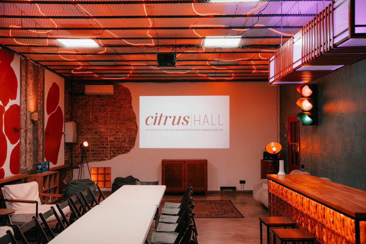 Арендовать Citrus Hall (Цитрус Холл). Зал Бергамот. Прямоугольное помещение с белым столом посередине, вокруг черные стулья. Левая стена с красным абстрактным рисунком, правая - серая. Потолок с сеткой и оранжевой неоновой подсветкой. Барная стойка справа, проектор с логотипом "citrus HALL" на дальней стене.