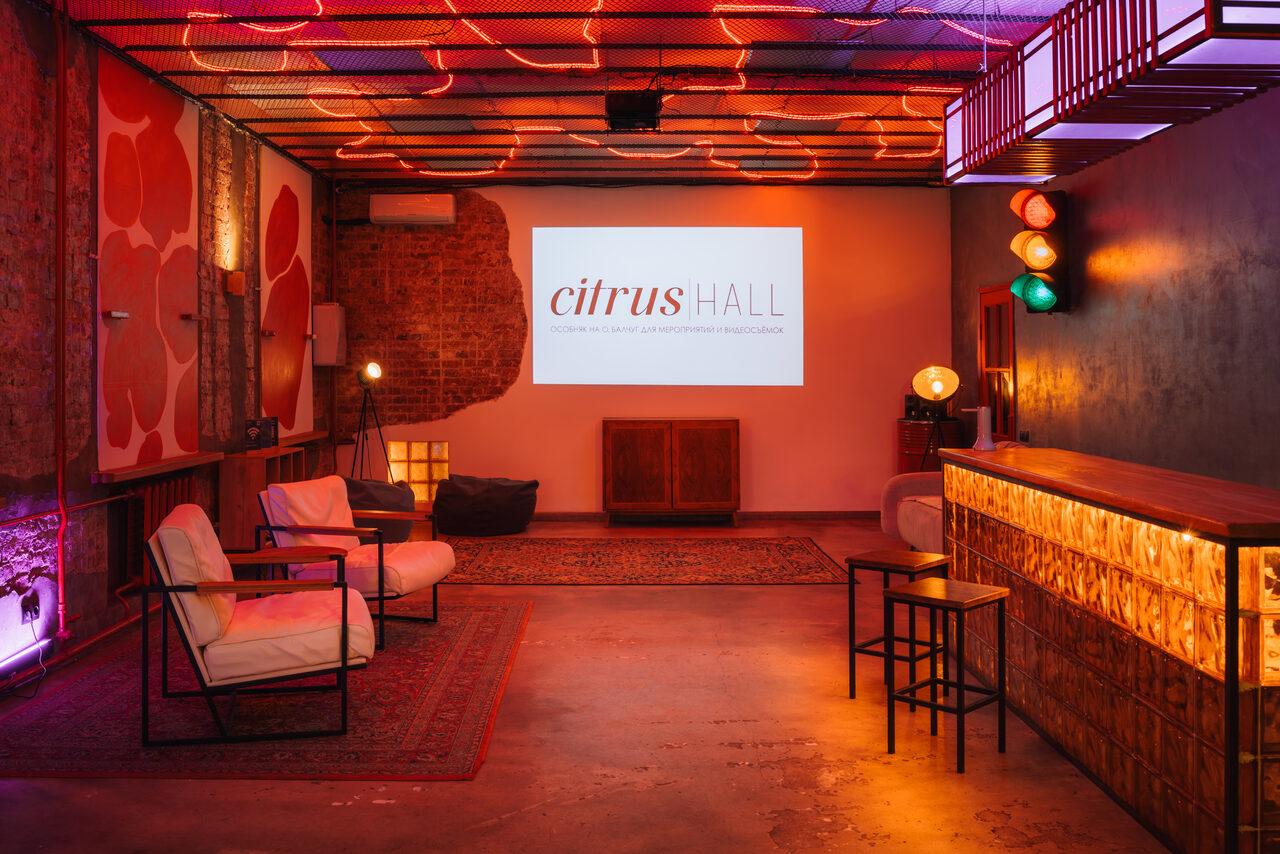 Арендовать Citrus Hall (Цитрус Холл). Зал Бергамот. Лофт-пространство с барной стойкой и креслами. Красный кирпич, бетонный пол, ковёр. Неоновое освещение на потолке и стенах. Стена с проектором, на котором текст: "citrus HALL". Лампа на штативе, мягкие пуфы, барные стулья.