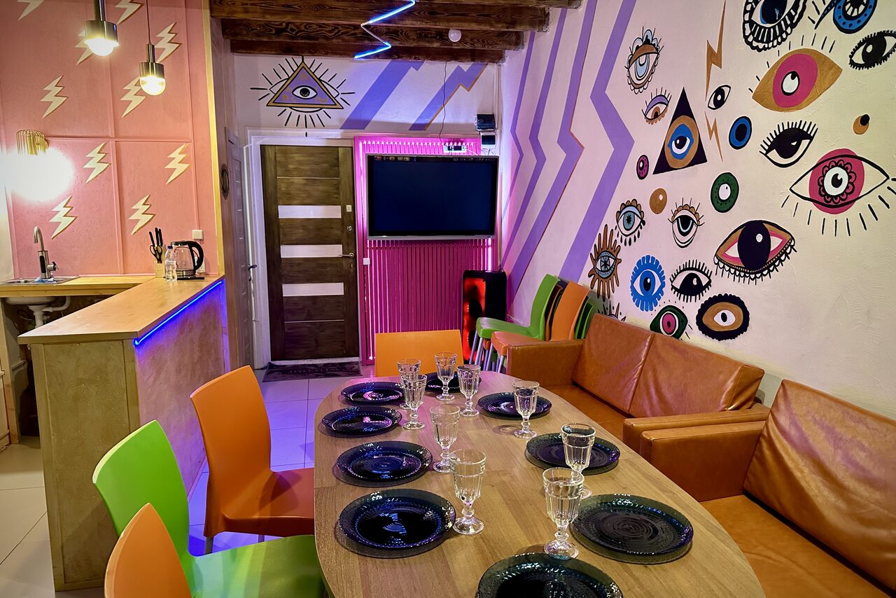 Арендовать Яркий двухуровневый лофт. Накрытый посудой стол. Разноцветные стулья. Диваны. Барная стойка. Разукрашенные рисунками стены разноцветные, яркие стены. Телевизор