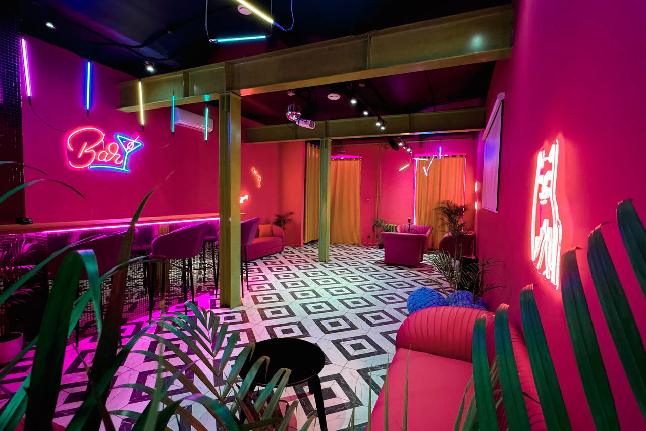 Арендовать Плэйс Лофт. Prisma Loft(Призма Лофт). Коктейль-бар с ярко-розовыми стенами, черно-белая плиточная мозаика на полу. Барная стойка с высокими стульями, неоновые вывески, разнообразные светильники. По периметру установлены диваны, интерьер дополняют растения.