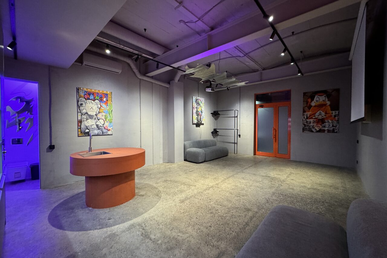 Арендовать Дизайнерский лофт-зал. Лофт-пространство с серыми бетонными стенами и полом, центральным круглым оранжевым столом, мягкими серыми диванами вдоль стен. Потолочное трековое освещение, несколько ярких картин, стеклянная дверь с оранжевой рамой.