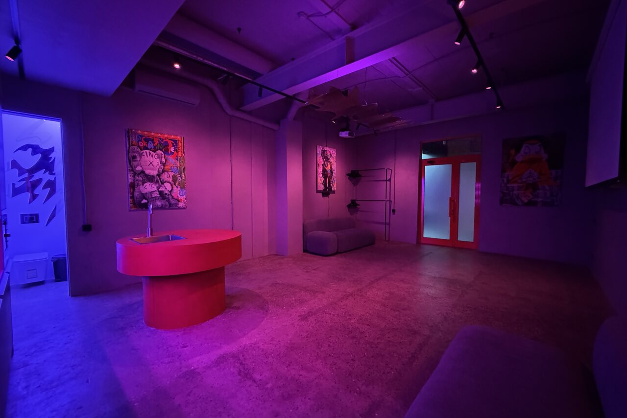 Арендовать Дизайнерский лофт-зал. Лофт-пространство с бетонными стенами и полом, мебель включает красный стол и серые диваны. На стенах картины, неоновые огни и фиолетовое освещение. На заднем плане стеклянные двери с красными рамами, открытая проводка на потолке.