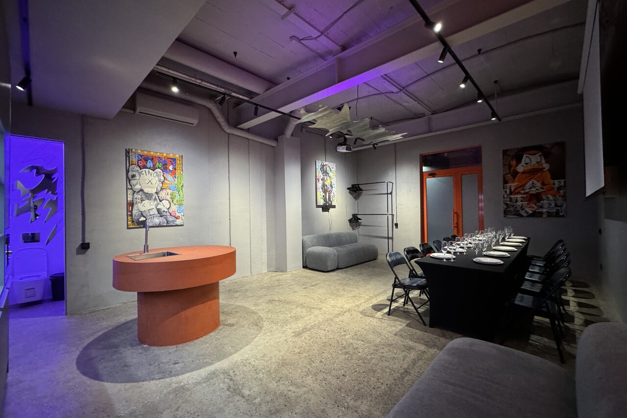 Арендовать Дизайнерский лофт-зал. Современное пространство с бетонными стенами и потолком, яркое освещение, круглый стол и стулья, длинный стол с сервировкой у стены, декоративные картины, серые мягкие диваны, красная дверь, лофт-стиль.
