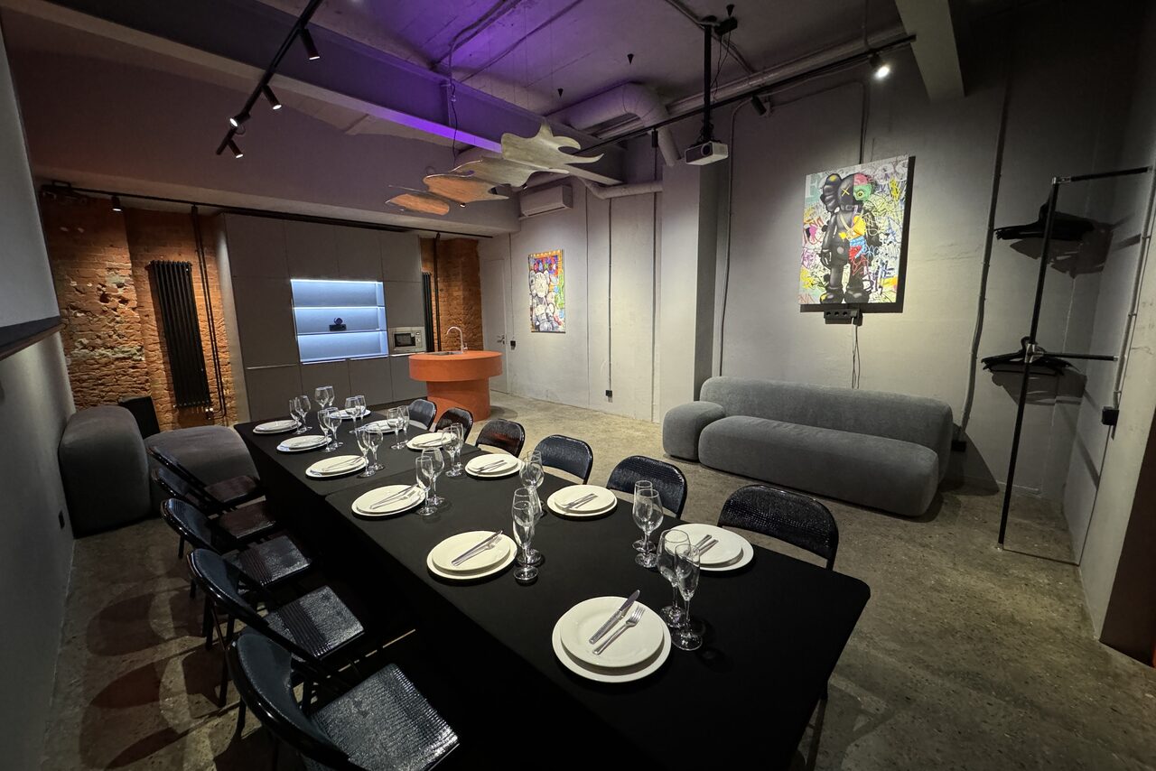 Арендовать Дизайнерский лофт-зал. Конференц-зал с длинным столом, накрытым черной скатертью, вокруг него стулья. Справа серый диван. На стенах несколько ярких картин. Освещение точечное и неоновое. Красный кирпич и серые стены. Потолок серый с трубами.