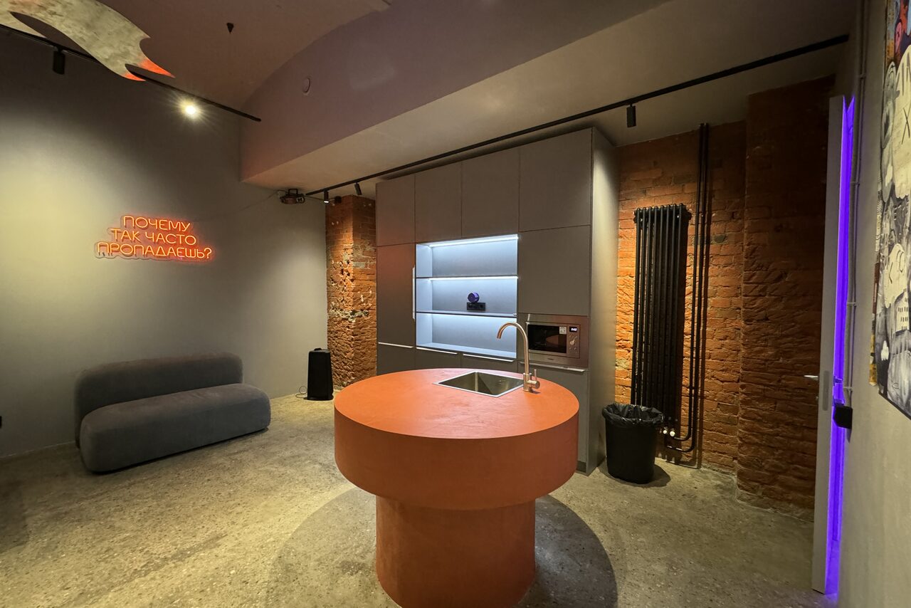 Арендовать Дизайнерский лофт-зал. Лофт-пространство с открытой кирпичной кладкой, круглым оранжевым столом с раковиной, серым диваном, настенным неоновым знаком, полки с подсветкой, металлическим радиатором и бетонным полом. Светлые и темные оттенки серого и оранжевого.