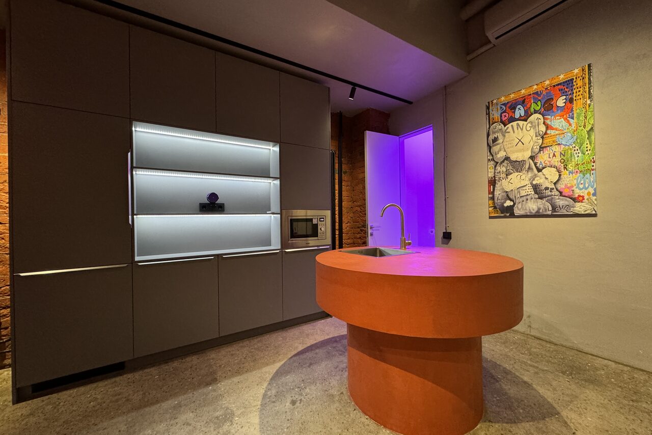 Арендовать Дизайнерский лофт-зал. Современное помещение с серыми встроенными шкафами и встроенной микроволновой печью. Центральный остров с круглым оранжевым столом и раковиной. На стене яркая картина. Светодиодное освещение, бетонный пол, открытая кирпичная кладка.