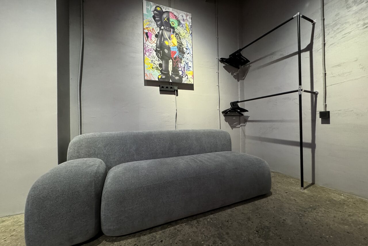 Арендовать Дизайнерский лофт-зал. Современное пространство с серым диваном, бетонным полом и светло-серыми стенами. На стене абстрактная картина. Справа металлическая стойка для одежды с вешалками. Освещение мягкое, рассеянное.