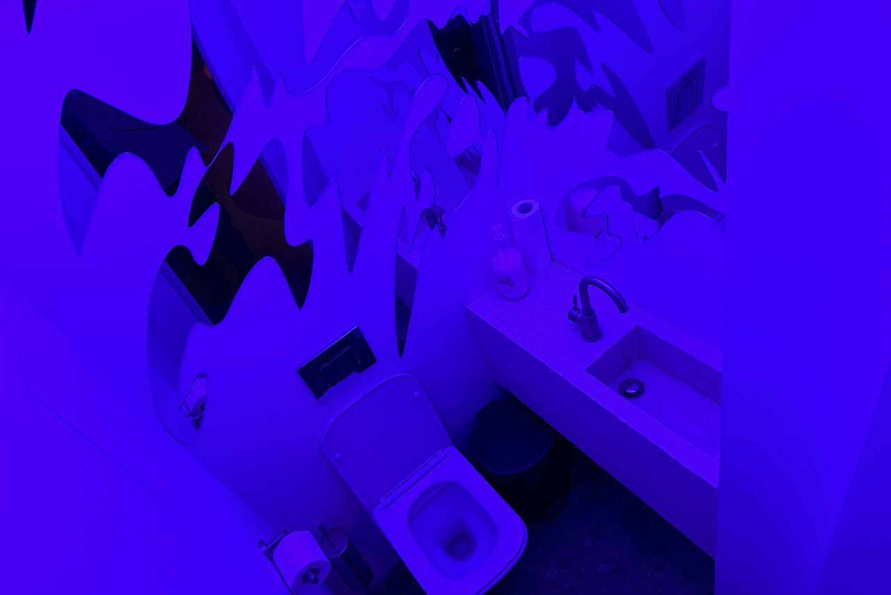 Арендовать Дизайнерский лофт-зал. Туалетная комната с унитазом, раковиной со смесителем, зеркалом, рулоном туалетной бумаги. Оформление в абстрактном стиле, яркое фиолетовое освещение. Стены и сантехника белые, пол темный.
