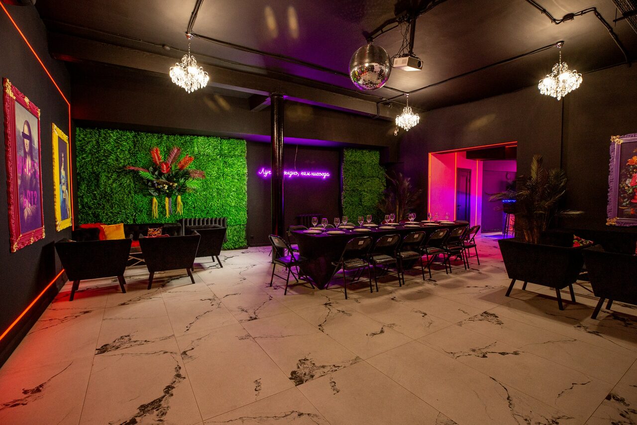 Арендовать Белый неоновый лофт-зал. Пространство с черными стенами и белым мраморным полом, декорировано зелеными растениями и неоновыми надписями. Столы и стулья черного цвета. Люстры и диско-шар на потолке. Яркое разноцветное освещение. На стенах картины.