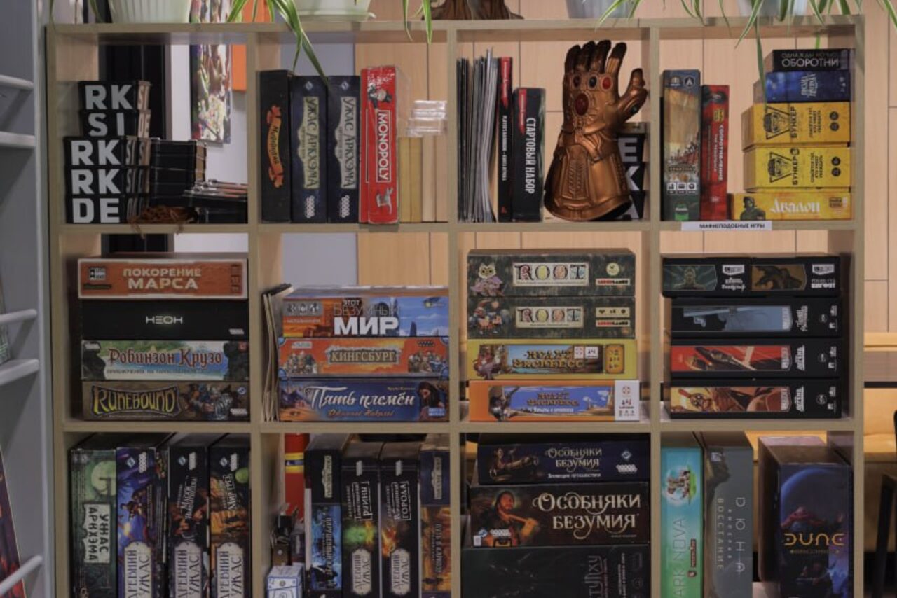 Арендовать Антикафе площадью 60 м² до 30 гостей. Стеллаж с настольными играми, разделенный на ячейки. В каждой ячейке коробки с играми разных размеров и цветов. В одной из ячеек фигурка в виде руки. Стеллаж из светлого дерева, хорошо освещён. Остальной интерьер не виден.