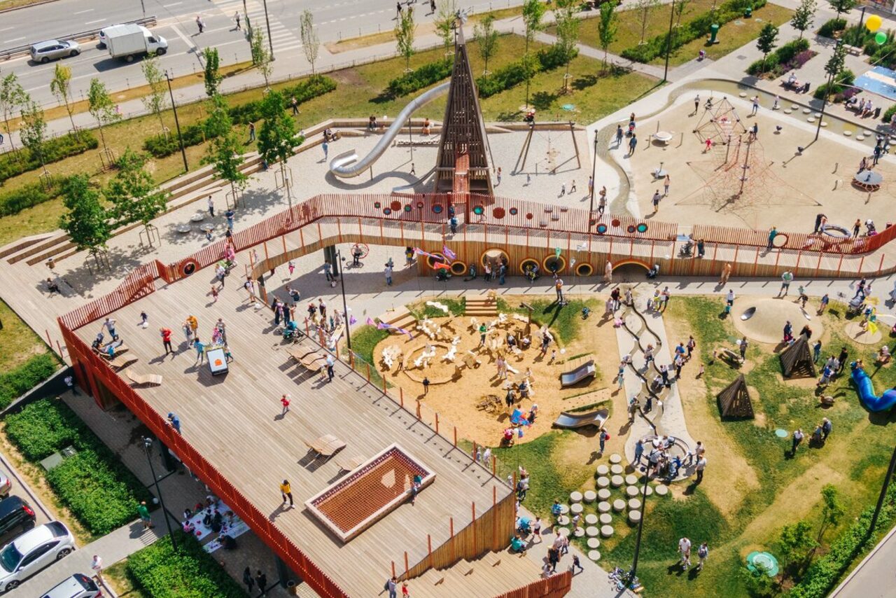 Арендовать Площадка в Москве c одной фотографией. Детская площадка с песочницей и игровыми зонами, окруженная деревянным настилом и забором. Присутствуют горки и спортивные конструкции. Растительность и прогулочные дорожки, светлое окружение, множество людей.