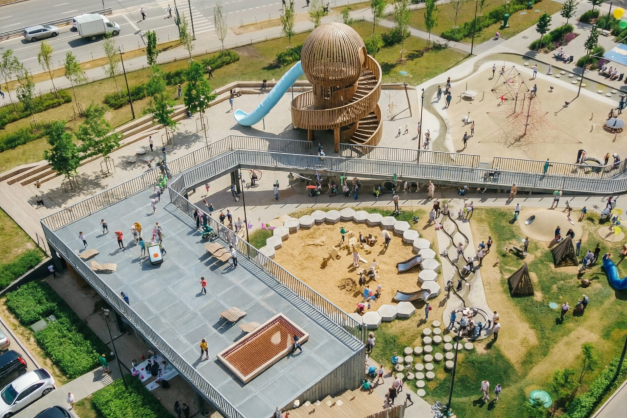 Арендовать Площадка в Москве c одной фотографией. Детская площадка с башней и спиральной лестницей, голубая горка. Песочница окружена скамейками. Асфальтовые и деревянные дорожки. Зелёные насаждения. Деревянные и верёвочные конструкции для лазания. Люди и дети гуляют и играют.