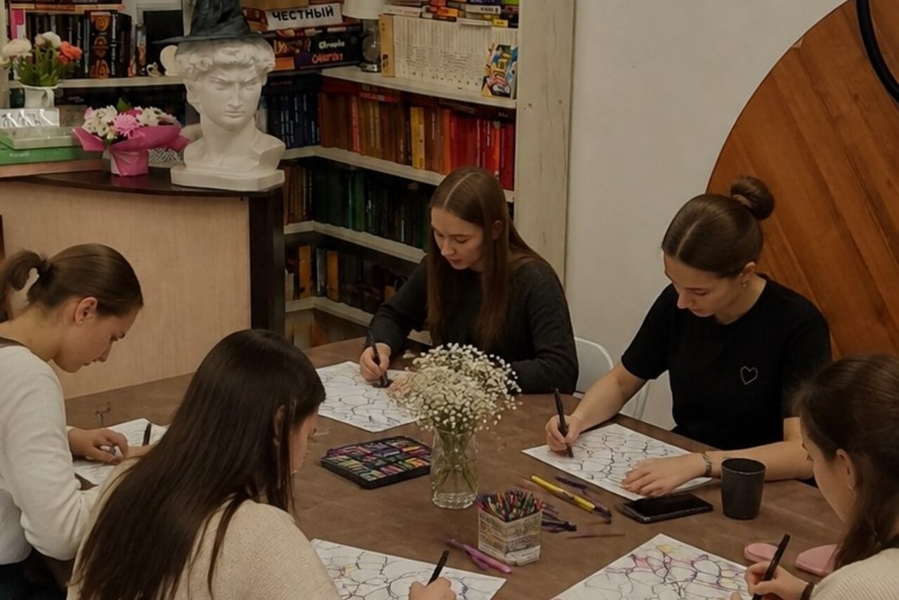 Арендовать Творческая лофт-студия под встречи и мастер-классы. Женщины сидят за столом в комнате с деревянными элементами и книжными полками. Стол украшен вазой с цветами, на нем лежат альбомы и карандаши. На заднем плане бюст и разные книги. Освещение мягкое, стены светлые.