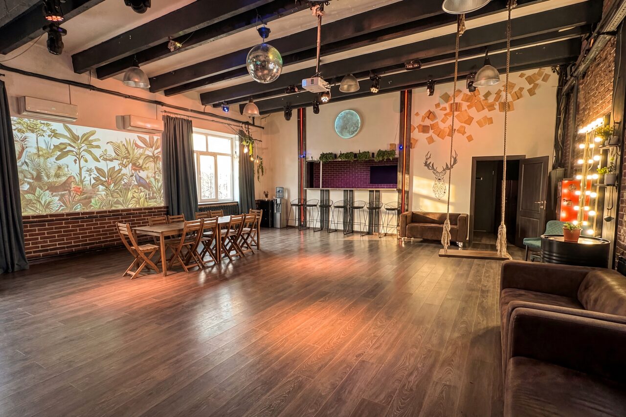 Арендовать Лофт с верандой, мангалом и сценическим оборудованием. Большое просторное помещение с деревянным полом. Стены из кирпича и штукатурки, частично декорированы рисунками листьев. Мебель: деревянный стол и стулья, барная стойка с высокими стульями, два дивана. Светильники подвесные, зеркальная шаровая лампа.