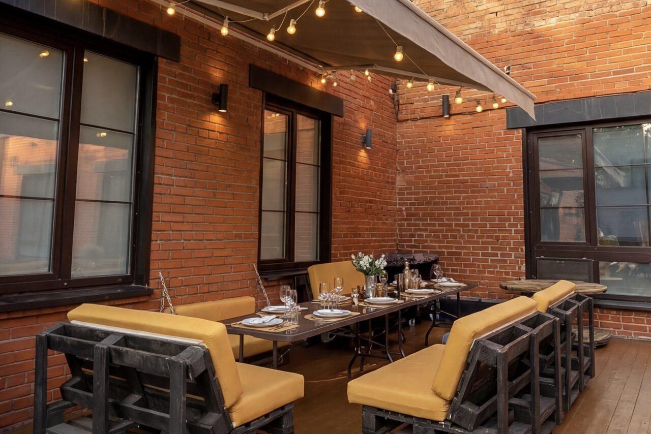 Арендовать Лофт с верандой, мангалом и сценическим оборудованием. Открытая терраса с кирпичными стенами и деревянным полом, стол для шести человек, обставленный приборами и украшенный вазой с цветами. Желтые подушки на лавках. Над столом натянут тент, развешены гирлянды с лампами.