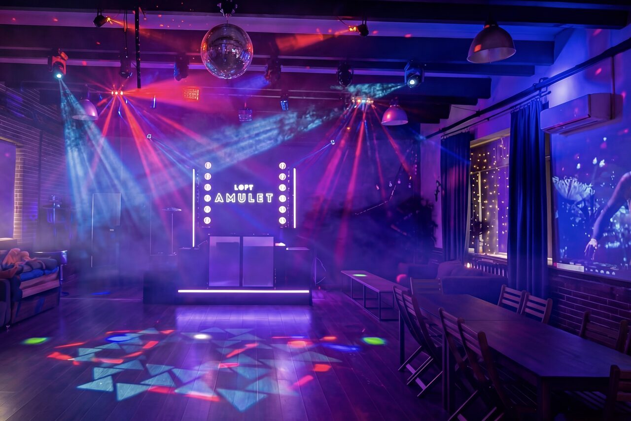 Арендовать Лофт с верандой, мангалом и сценическим оборудованием. Loft-пространство с деревянным полом и кирпичными стенами. В центре сцена с подсветкой и надписью. Расставлены столы и стулья. Дым и световые проекции от диско-шара. Яркое разноцветное освещение. Текст на стене: "Loft Amulet".