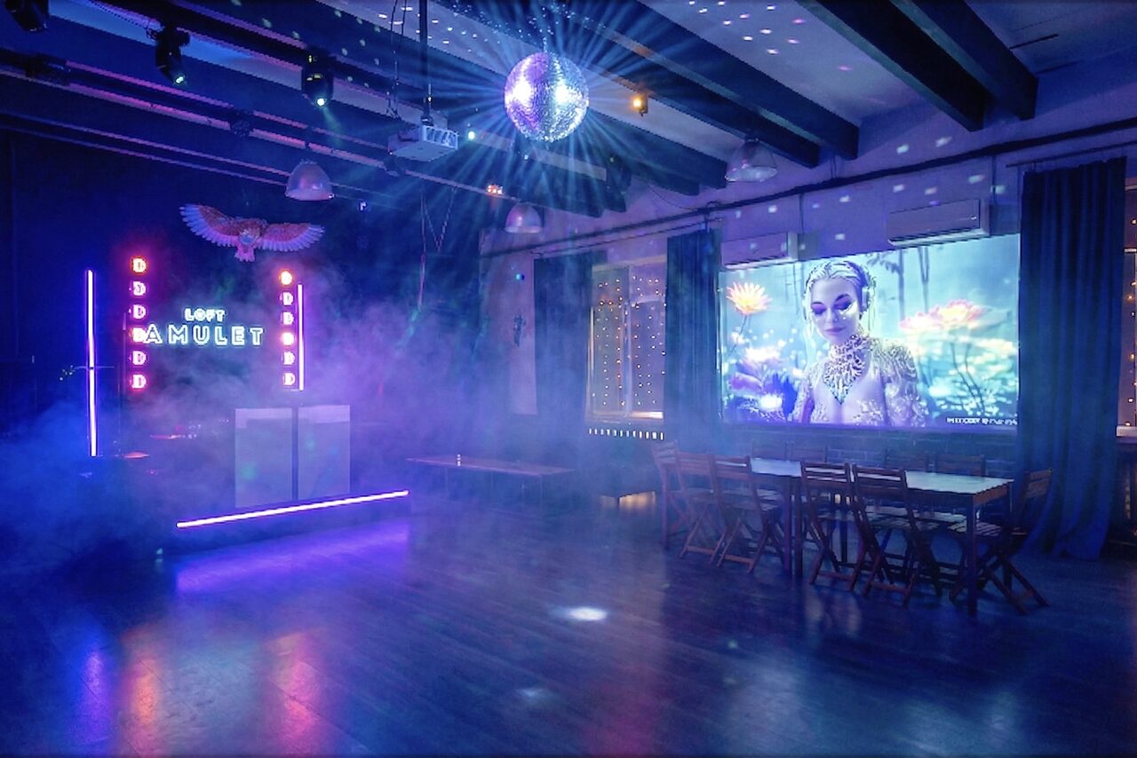 Арендовать Лофт с верандой, мангалом и сценическим оборудованием. Пространство с деревянным полом, столами и стульями. Неоновая вывеска с надписью "LOFT", зеркальный шар на потолке. Проекция изображения на стене, фоновая подсветка и дым. Потолочные балки, тёмно-синие шторы.