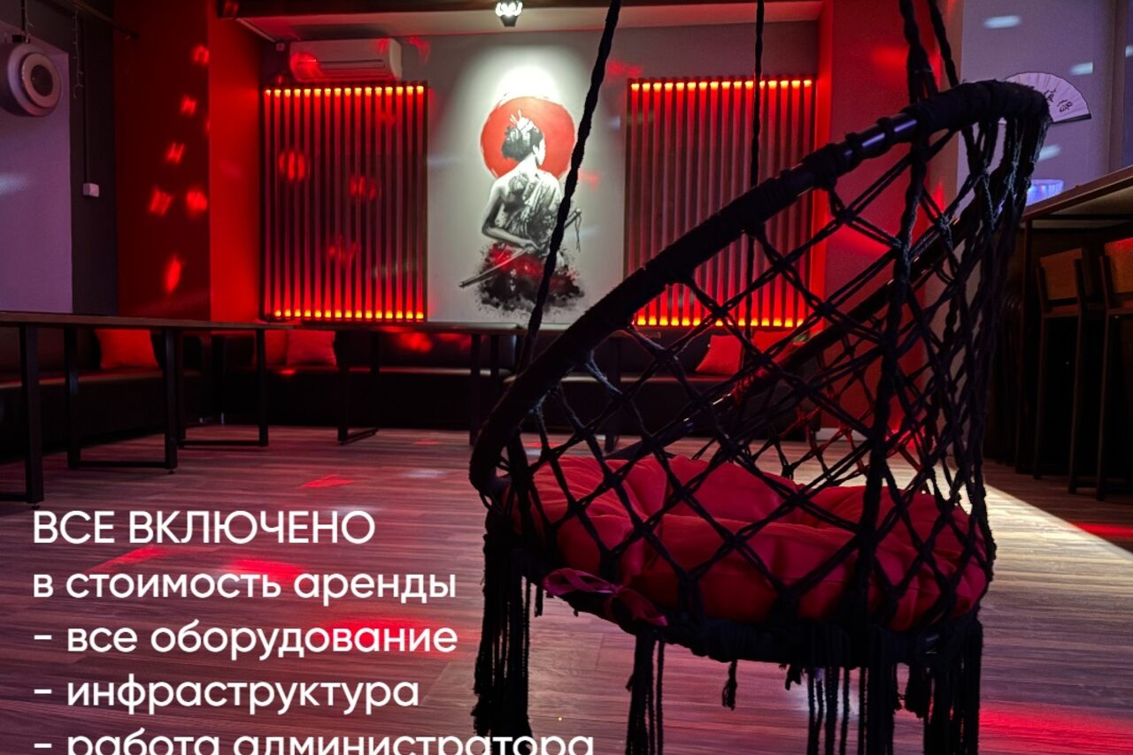 Арендовать Лофт ИЗНАНКА. Зал ТОКИО. Пространство с затемненным освещением, красные световые акценты на стенах. Мебель: черные скамьи вдоль стены, плетеное подвесное кресло с красной подушкой. На стене картина с изображением женщины. На полу паркет. Текст: "Все включено в стоимость аренды - все оборудование, инфраструктура".