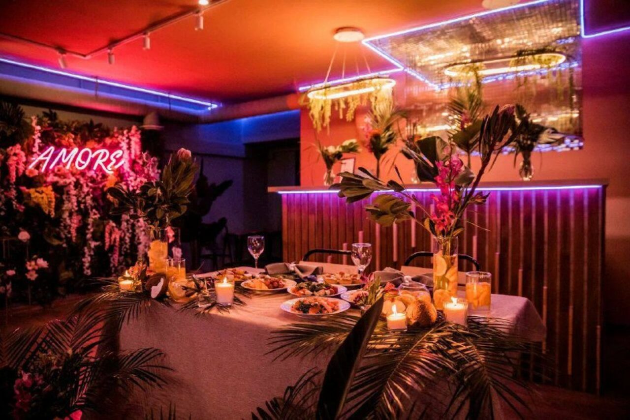 Арендовать Тропический лофт с современным дизайном и верандой. Пространство с обеденным столом, сервированным блюдами и напитками. Окружение украшают зелёные растения и свечи. На заднем плане фоновая неоновая вывеска «AMOR». Освещение разноцветное: красное, синее и тёплое, потолок отражает свет зеркальным материалом.