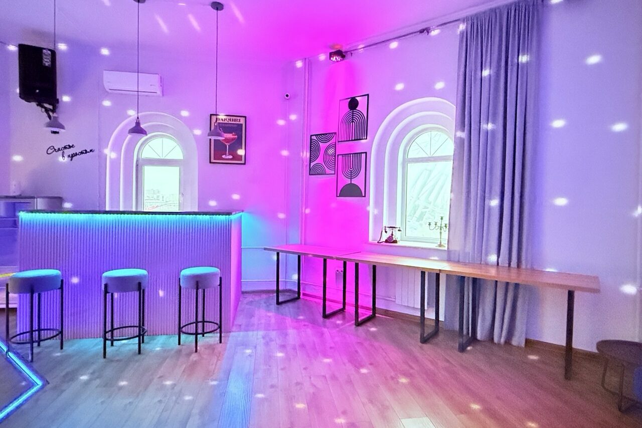 Арендовать Центрум. Уютное светлое пространство круглой формы. Просторная комната с барной стойкой слева, тремя высокими стульями, длинным деревянным столом справа. Арочные окна, занавески, светодиодная подсветка розового и синего цветов, паркетный пол, современные постеры на стенах.