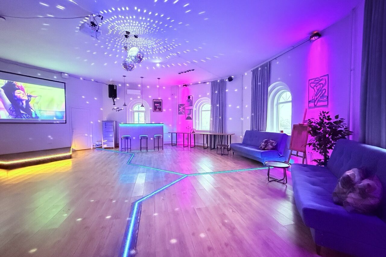 Арендовать Центрум. Уютное светлое пространство круглой формы. Просторное помещение с деревянным полом, несколькими диванами и барной стойкой. На стене экран с проектором. Праздничное освещение с диско-шаром и цветными светодиодами. Высокие окна с арками и занавесками. На стенах постеры и растения.