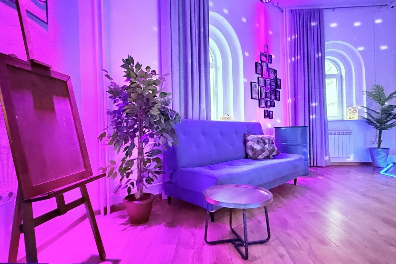 Арендовать Центрум. Уютное светлое пространство круглой формы. Просторная комната с деревянным полом, синим диваном и круглым столиком. На стене фотографии, светодиодная подсветка розового и голубого цветов. Растения в кадке и мольберт. Арочные окна с серыми шторами.