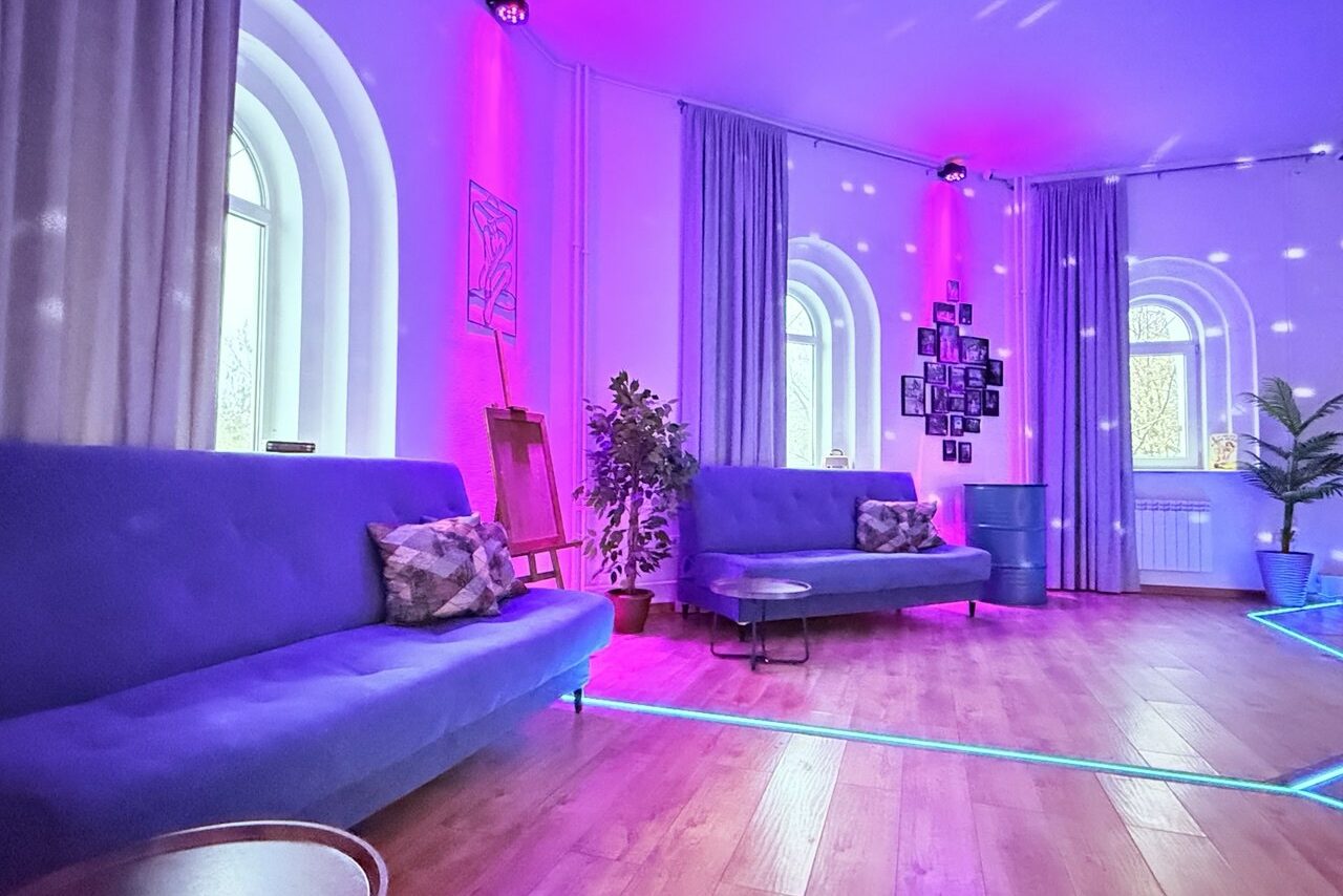 Арендовать Центрум. Уютное светлое пространство круглой формы. Большая комната с деревянным полом, двумя синими диванами, подушками, металлическими столиками и растениями. Окна арочные, стены украшают фотографии и картины. Освещение неоновое, фиолетово-розовое. На полу светодиодная подсветка.