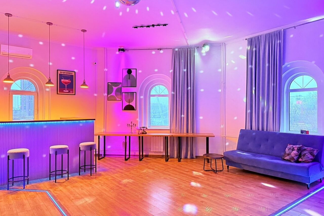 Арендовать Центрум. Уютное светлое пространство круглой формы. Просторное и светлое помещение с деревянным полом; барная стойка с тремя высокими стульями; рядом столы и синий диван; на стенах картины; арочные окна с серыми занавесками; разноцветное освещение и точечные светильники.