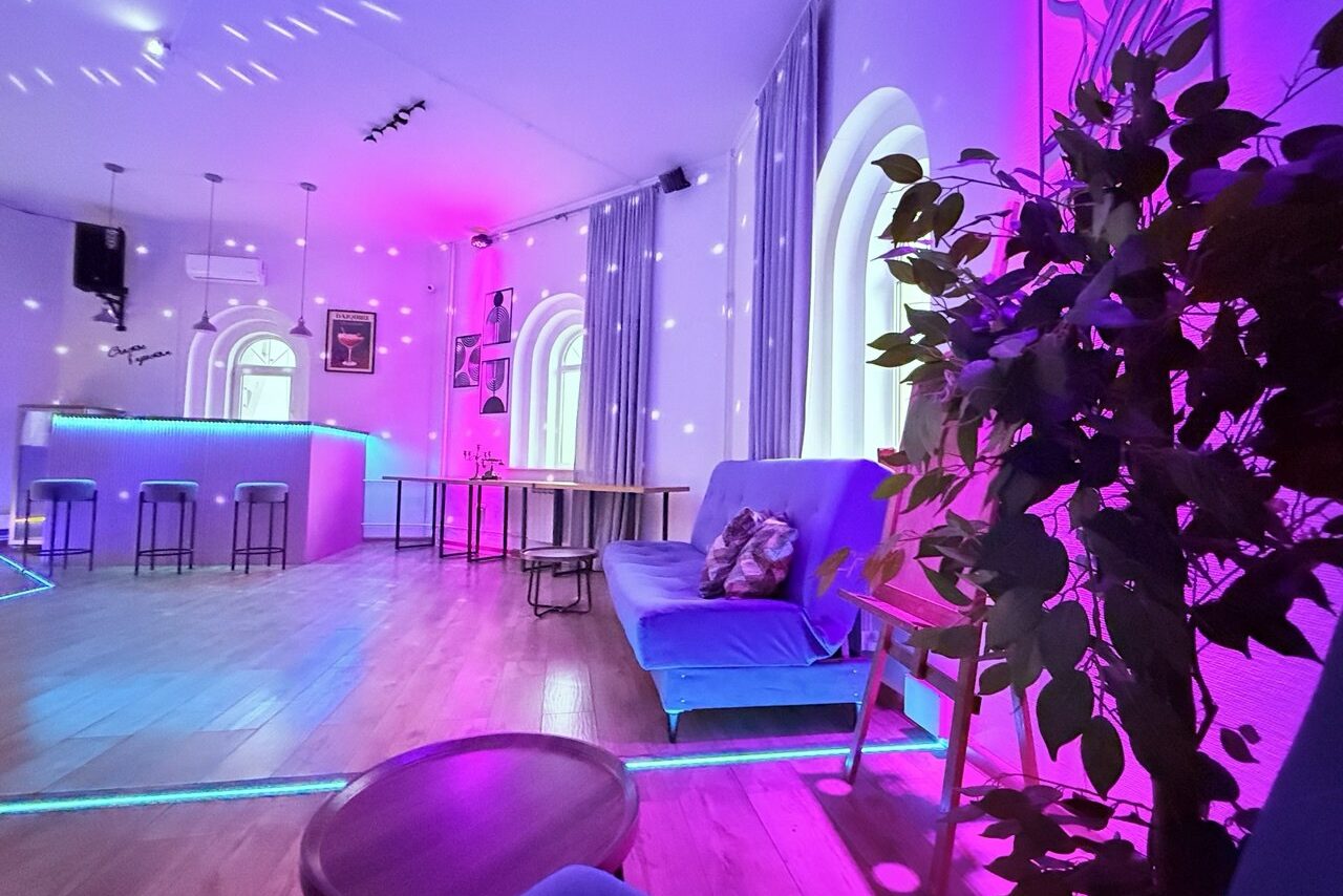 Арендовать Центрум. Уютное светлое пространство круглой формы. Просторная комната с барной стойкой, высокими табуретами и диваном. Окна арочные, пол деревянный. Освещение разноцветное, в основном пурпурное. На стенах и потолке световые эффекты. В углу растения, столы и стулья.