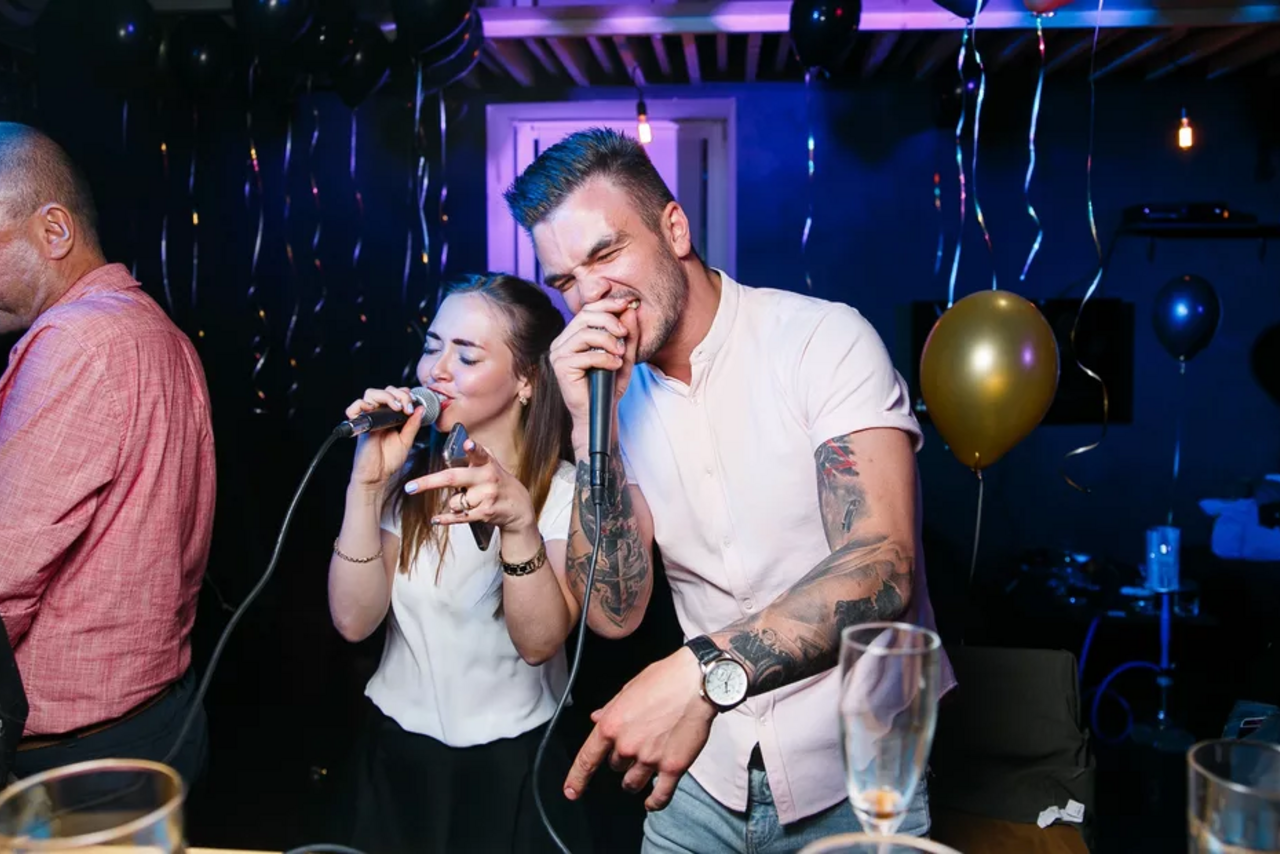 Арендовать Тестовый зал мск. Караоке-вечеринка в помещении с тёмными стенами, разноцветные воздушные шары, светодиодное освещение, деревянные балки на потолке, микрофоны, стеклянные бокалы на переднем плане, мужчина и женщина поют в микрофоны.