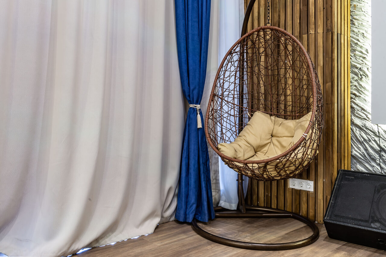 Арендовать Rubin Loft (Рубин Лофт). Зал Сицилия. Пространство с подвесным креслом-коконом на металлической стойке, обитым подушками. Стена с деревянными панелями и драпировкой из белой и синей ткани. На полу деревянное покрытие. Видна часть зеркала и чемодан.