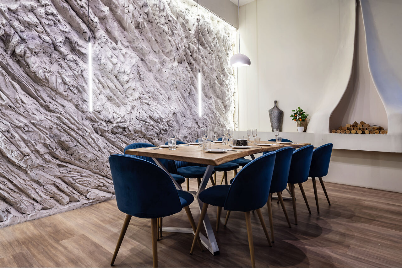 Арендовать Rubin Loft (Рубин Лофт). Зал Сицилия. Современное помещение с большим деревянным столом и синими стульями. Стена декорирована текстурной поверхностью в серых тонах, оборудована вертикальными светильниками. Светло-коричневый пол, декоративный камин с полкой и вазой.