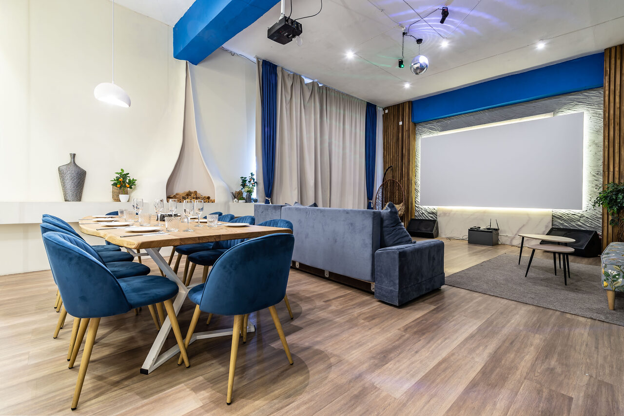 Арендовать Rubin Loft (Рубин Лофт). Зал Сицилия. Современное пространство с деревянным полом, обеденным столом и синими стульями. Серая мягкая мебель рядом с проектором и экраном. Потолочные светильники и декоративные элементы. Светлые шторы, зелёные растения на фоне.