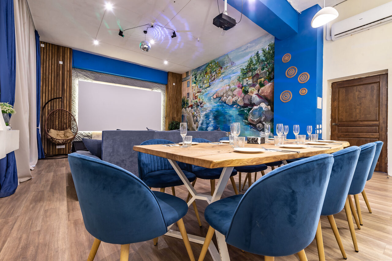 Арендовать Rubin Loft (Рубин Лофт). Зал Сицилия. Пространство для мероприятий с обеденным столом и синими стульями, деревянный пол, синий и белый декор, настенная роспись с пейзажем, потолочные светильники, подвесное кресло, деревянная дверь, проекционный экран и стеклянные бокалы на столе.