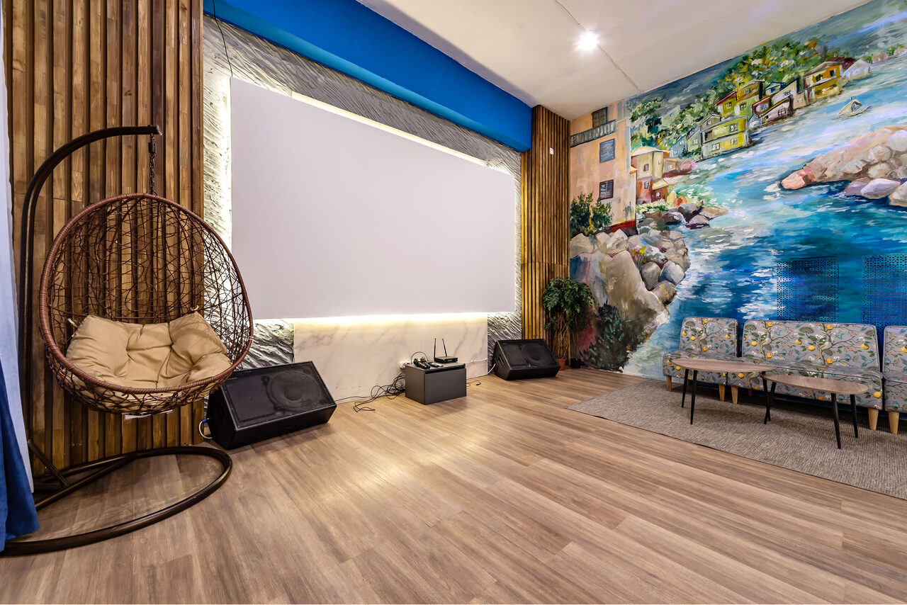 Арендовать Rubin Loft (Рубин Лофт). Зал Сицилия. Просторная комната с деревянным полом. На стене роспись с пейзажем. Имеется проектор, пара колонок, висящее кресло-кокон, пара стульев с серой обивкой и растительность. Стена оформлена под дерево, освещение точечное и диодное.