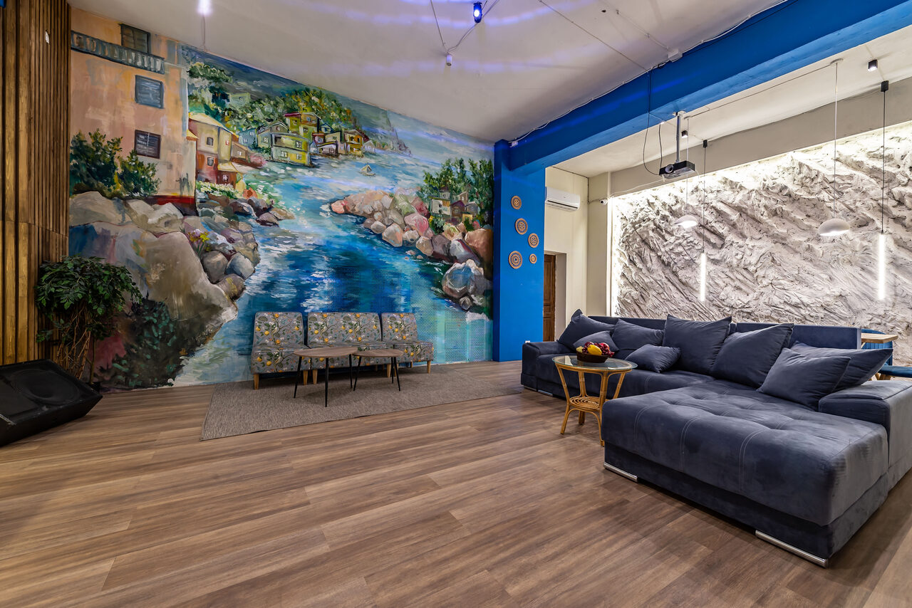 Арендовать Rubin Loft (Рубин Лофт). Зал Сицилия. Просторная комната с деревянным полом, синяя стена с настенной росписью на тему реки и домов. Серый угловой диван с плетеным столиком, напротив — три стула. Современное освещение, декоративные элементы на стенах.
