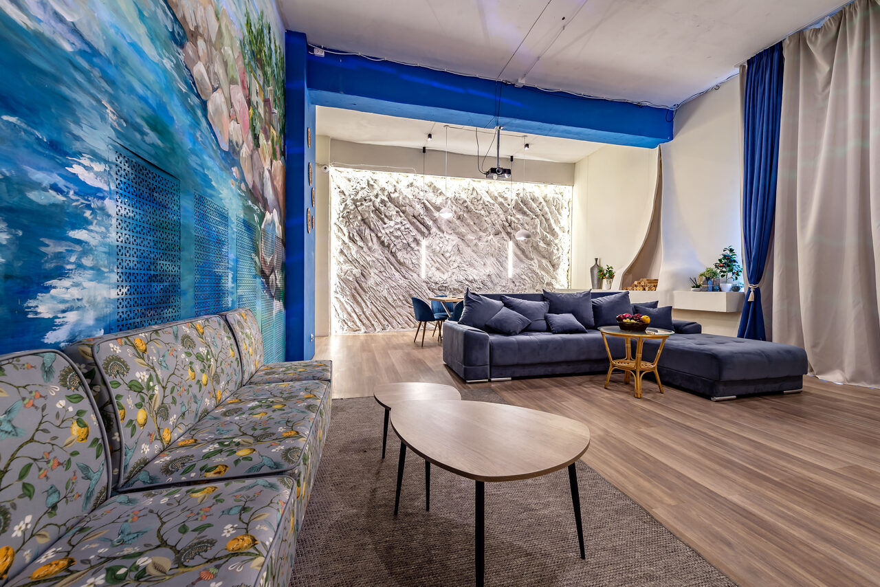 Арендовать Rubin Loft (Рубин Лофт). Зал Сицилия. Просторное помещение с диванами, обеденной зоной и стульями. Стены отделаны текстурированными панелями и росписью. Потолочные светильники. Пол деревянный, на полу коврики. Шторы темно-синие, декоративная зелень.