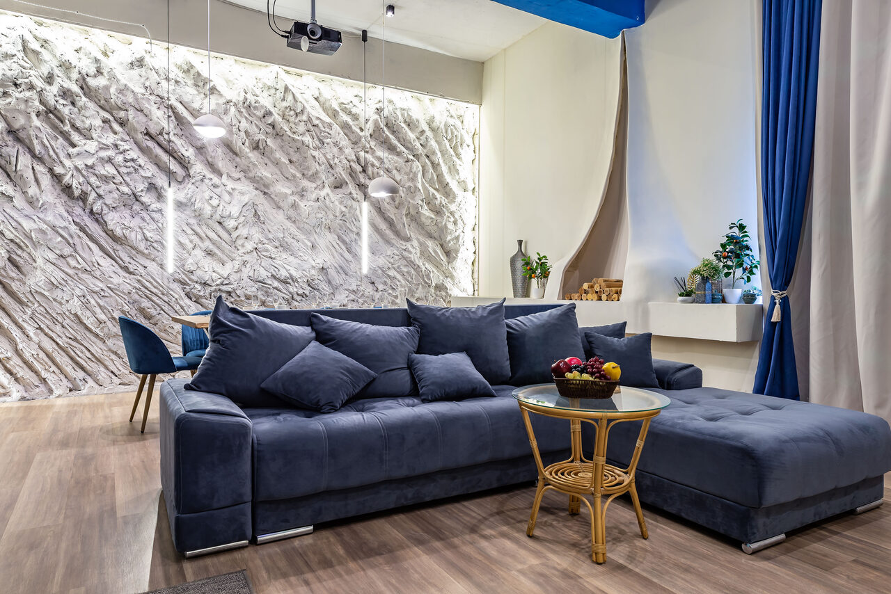 Арендовать Rubin Loft (Рубин Лофт). Зал Сицилия. Современное пространство с серым угловым диваном, круглым стеклянным столиком на деревянных ножках, декоративной стеной с подсветкой, подвесными светильниками, синими шторами, деревянным полом и белыми стенами.