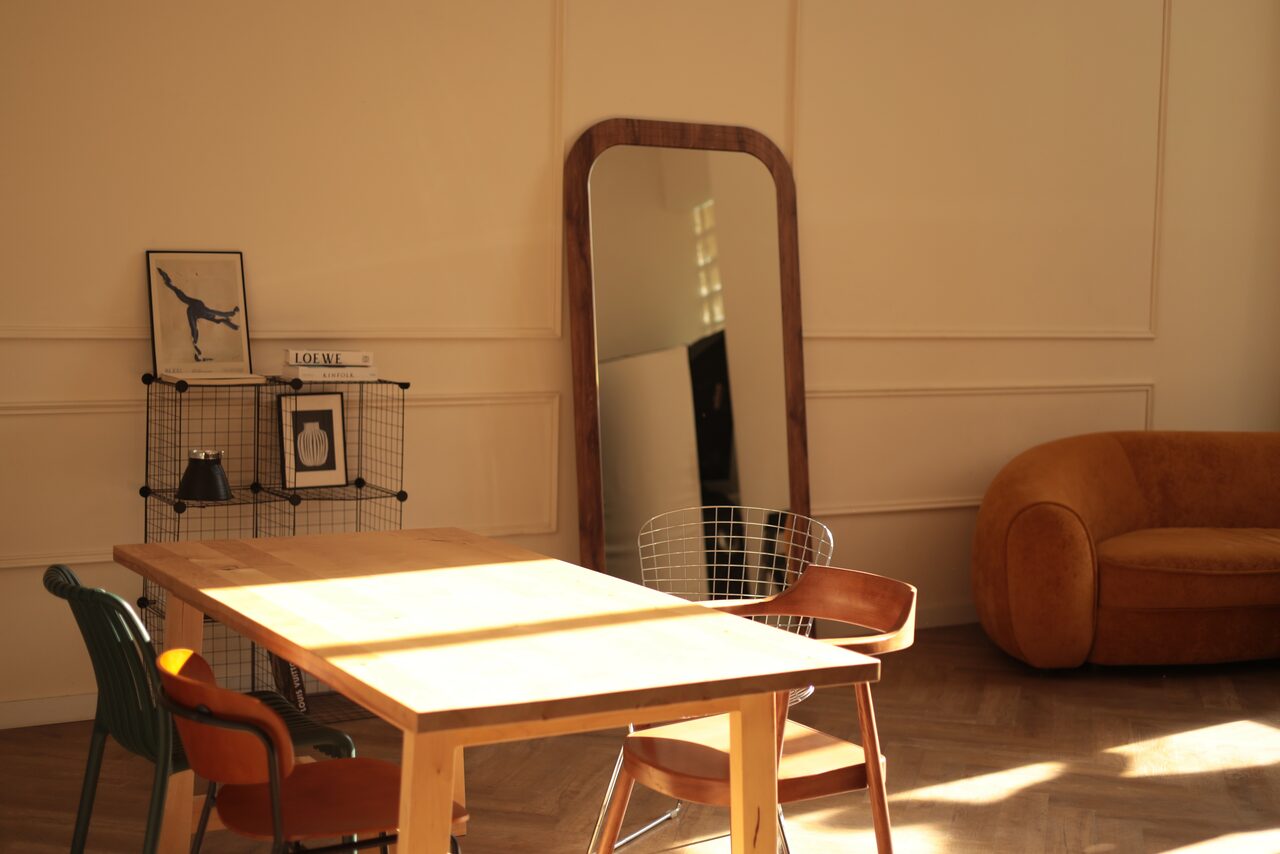 Арендовать Стильный лофт для уютных вечеринок. Светлая комната с деревянным полом и панельными стенами. Большое зеркало в деревянной раме стоит у стены. Деревянный стол окружён разноцветными стульями. Металлическая полка с книгами и фотографиями. Справа мягкий оранжевый диван. Естественное освещение.