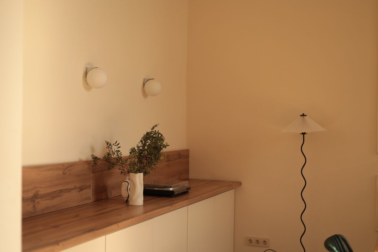 Арендовать Стильный лофт для уютных вечеринок. Комната с бежевыми стенами и деревянной отделкой. На столешнице белая ваза с зеленью и ноутбук. На стене два круглых светильника. Справа лампа с изогнутой ножкой и круглым абажуром. Белые шкафы внизу.