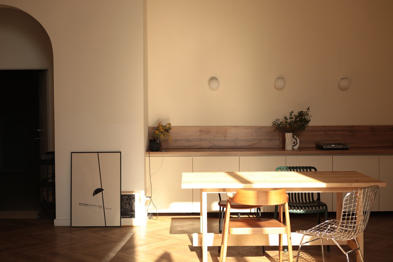 Арендовать Стильный лофт для уютных вечеринок. Пространство с деревянной обеденной мебелью, длинным столом и разнообразными стульями. Светлые стены, паркетный пол, большое арочное окно в тени. На стене несколько современных настенных светильников. На тумбе декор и комнатные растения.