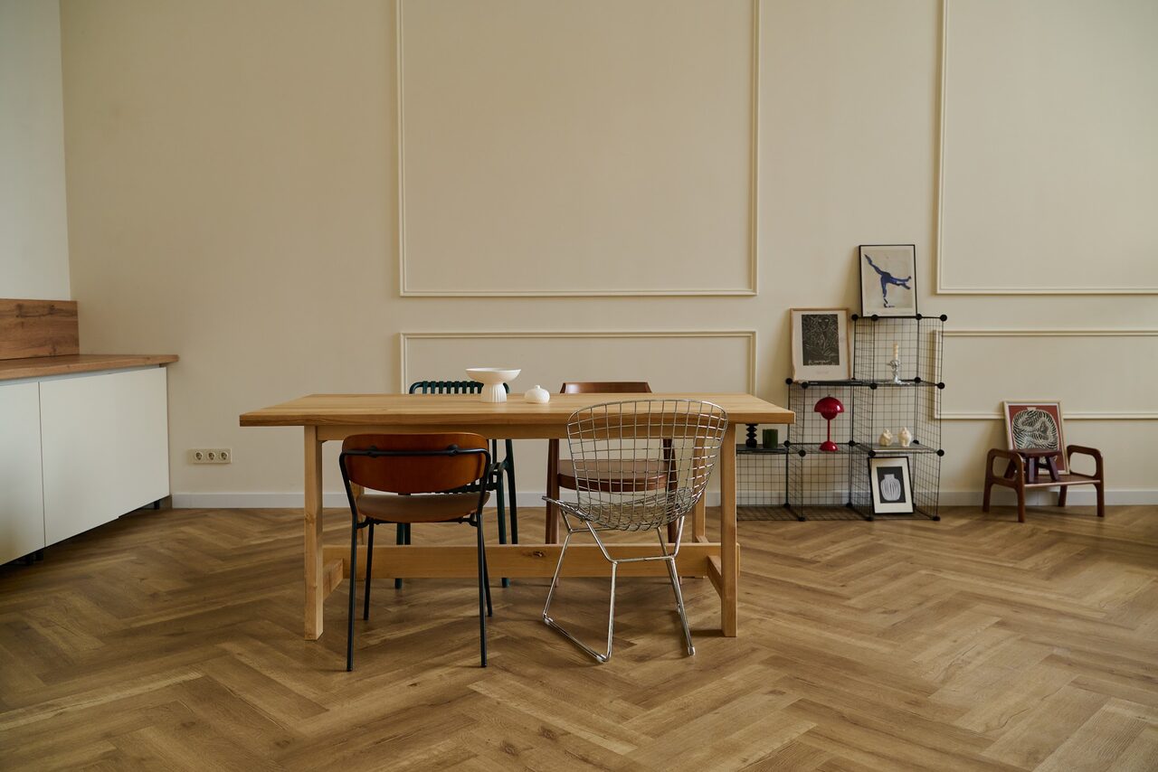 Арендовать Стильный лофт для уютных вечеринок. Современное помещение с деревянным полом елочкой, светлыми стенами с лепниной. Деревянный стол с четырьмя различными стульями. Слева белые шкафы, справа металлический стеллаж с декором, рядом картина и кресло. Естественное освещение.