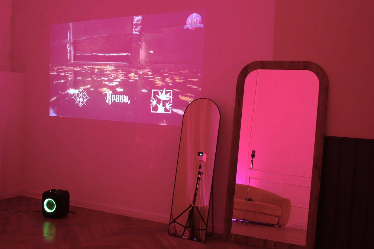 Арендовать Стильный лофт для уютных вечеринок. Комната с проектором на стене, показывающим красочное изображение. На полу черная колонка. Два больших зеркала, одно с деревянной рамой, другое на треноге. Слабое розовое освещение, диван виден в отражении зеркала. Паркетный пол.