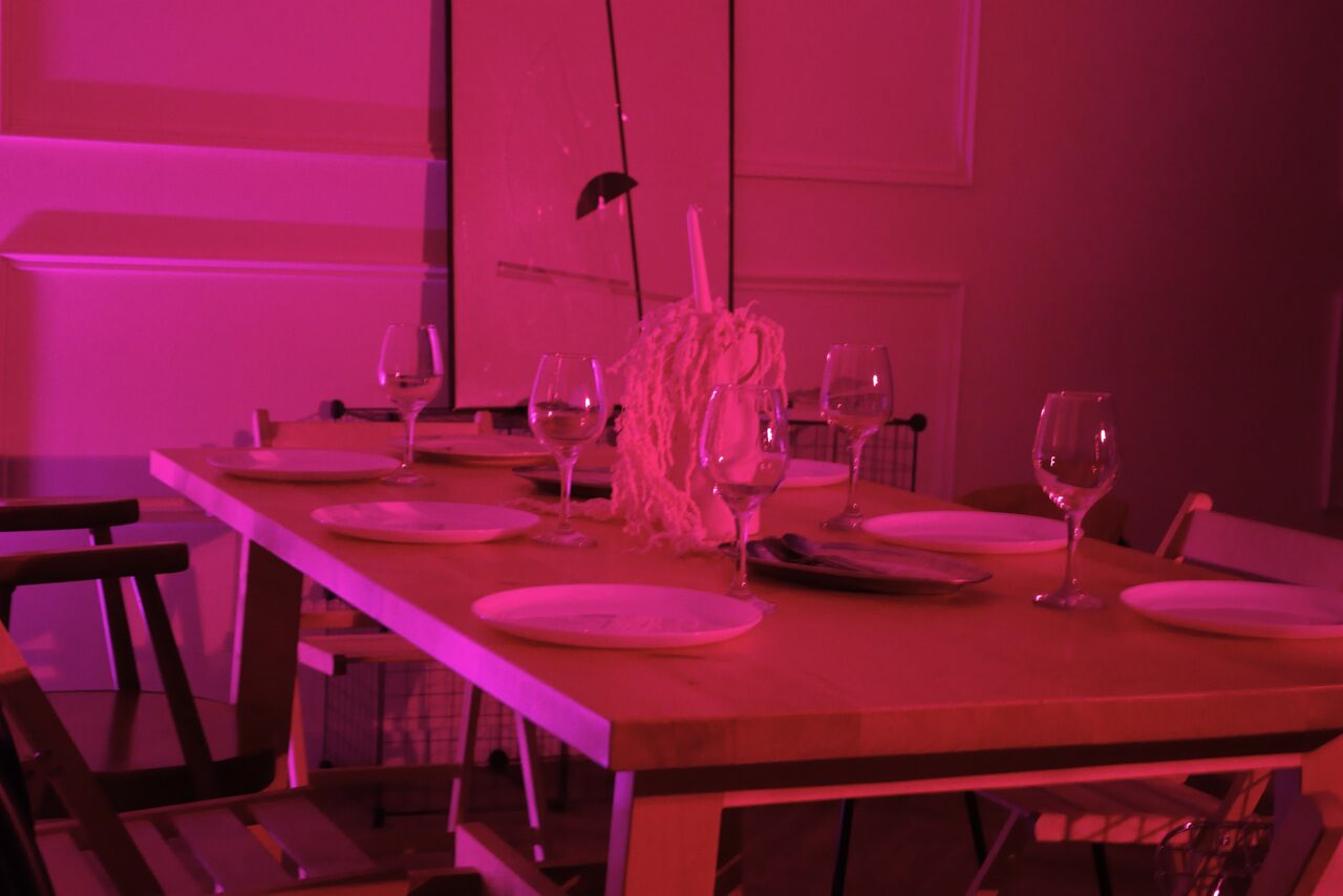 Арендовать Стильный лофт для уютных вечеринок. Столовая в розовом освещении, деревянный стол с шестью накрытыми приборами, бокалы для вина, декоративная композиция по центру. Стулья разных дизайнов вокруг стола. Белые наклонные панели на стенах.