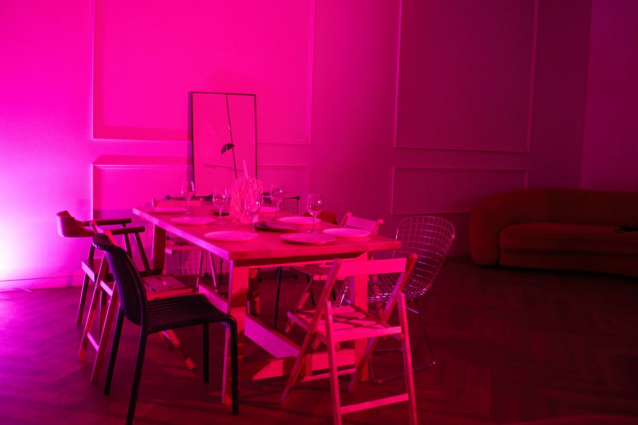 Арендовать Стильный лофт для уютных вечеринок. Просторная комната с деревянным полом, розовым освещением. В центре деревянный стол с посудой и бокалами, окруженный разноцветными стульями. Слева зеркало, справа коричневый диван. Стены украшены панельным декором.