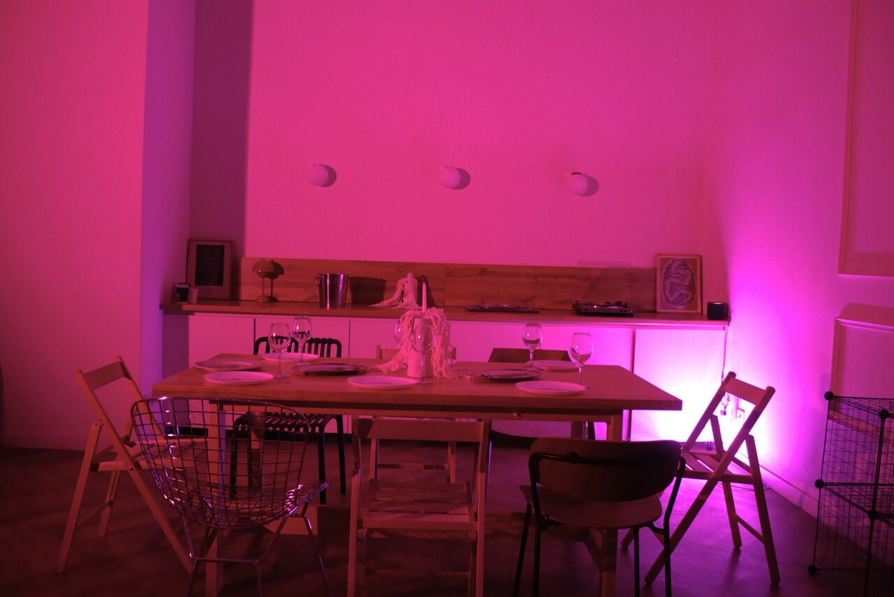 Арендовать Стильный лофт для уютных вечеринок. Столовая зона с деревянным столом, окруженным разными стульями, на столе тарелки и бокалы. На заднем плане открытые полки с предметами и декоративное освещение розового цвета. Белые стены, паркетный пол. Трековые светильники на стене.