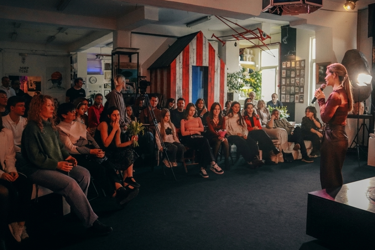 Арендовать А53: Secret Gallery (Секретная галерея). Просторное помещение с темным ковровым покрытием, кресла и стулья для зрителей, выступающая женщина с микрофоном, декоративный фоновый элемент в виде домика с красными и синими полосами, боковое освещение, растения и картины на стенах.